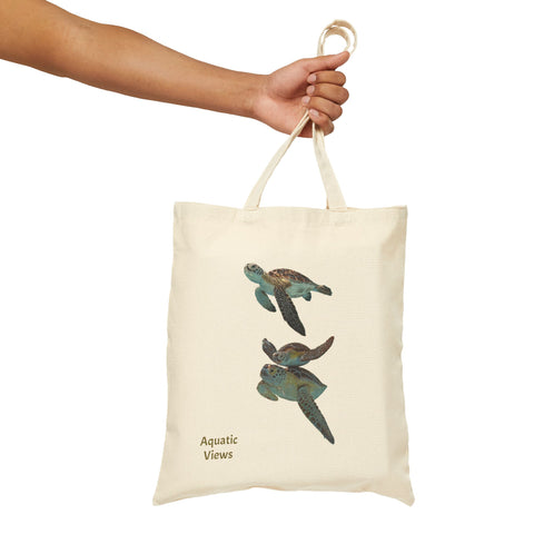 Hand holding a natural cotton canvas tote bag with a sea turtle print and 'Aquatic Views' text. Lightweight and durable.