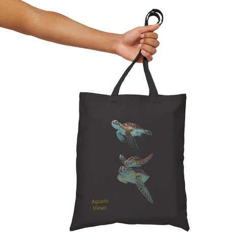 Hand holding a black cotton canvas tote bag featuring a sea turtle print and 'Aquatic Views' text. Durable and versatile.